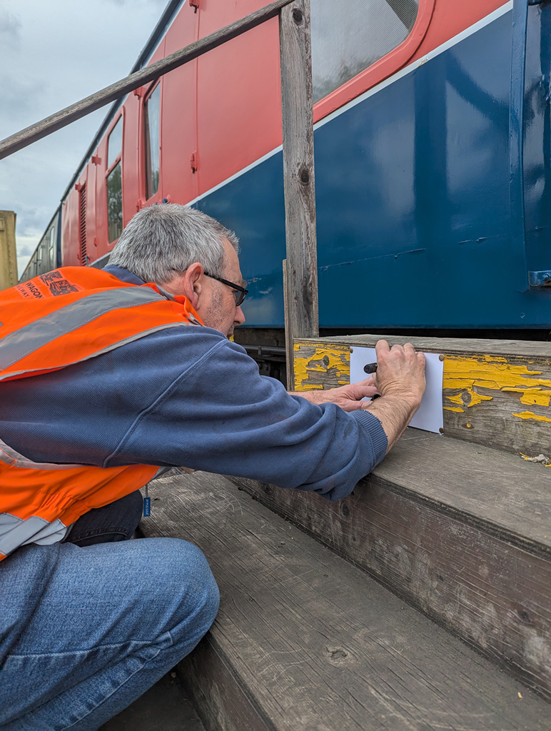 A person wearing an orange safety vest is crouched down, using a marker to make notes on a piece of paper attached to a wooden step next to a railway carriage.