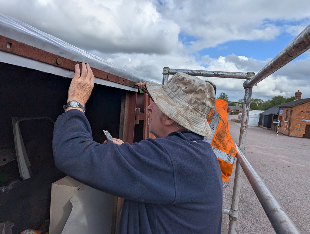 A person on a scaffold is inspecting the edge of a structure, wearing a hat and a blue sweater, with some tools in hand.