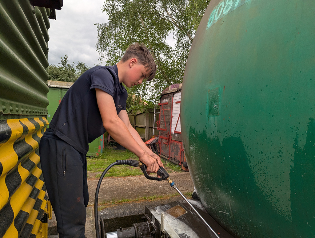 A young person is using a pressure washer to clean a large green tank, while standing next to a yellow and black striped structure in a yard.