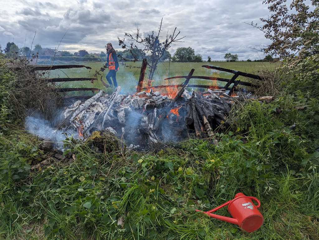A person in an orange vest walking near a controlled fire, surrounded by smoke and greenery, with a red watering can lying on the ground.